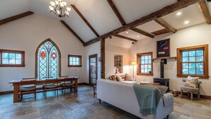 Une pièce spacieuse avec un plafond haut, des poutres apparentes, une grande fenêtre à vitraux, une cheminée et un coin salon confortable.