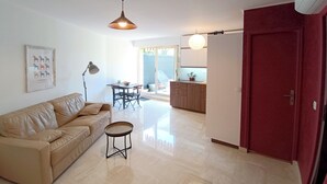 Sala de estar com sofá, mesa redonda pequena, luminária pendente e área de cozinha com armários e uma porta vermelha.