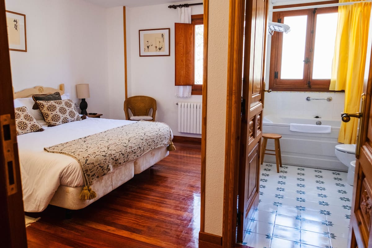 A bedroom with a bed, a chair, a window, and a bathroom with a bathtub and toilet.
