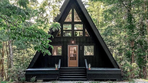 Une cabane noire avec un toit triangulaire, une porte d’entrée et un petit porche avec des marches.