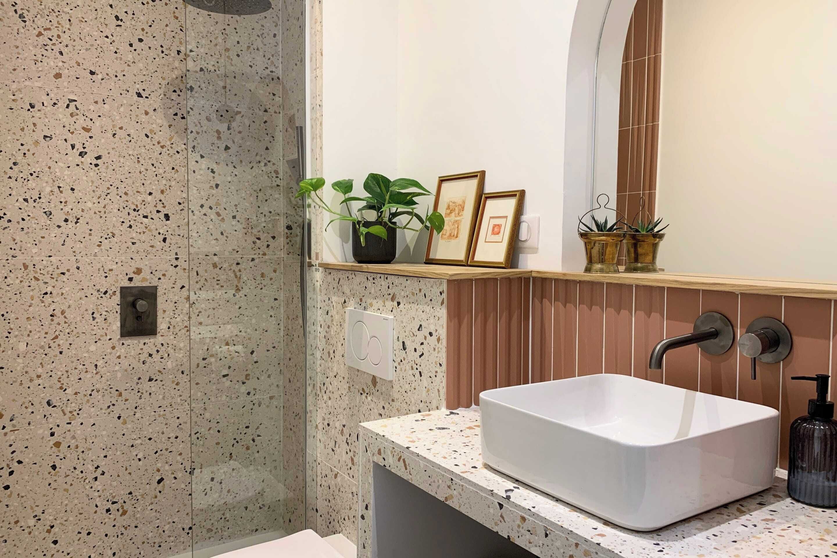 A bathroom with a speckled tile wall, a white sink with a modern faucet, and a shelf with potted plants and framed pictures.