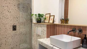 A bathroom with a speckled tile wall, a white sink with a modern faucet, and a shelf with potted plants and framed pictures.