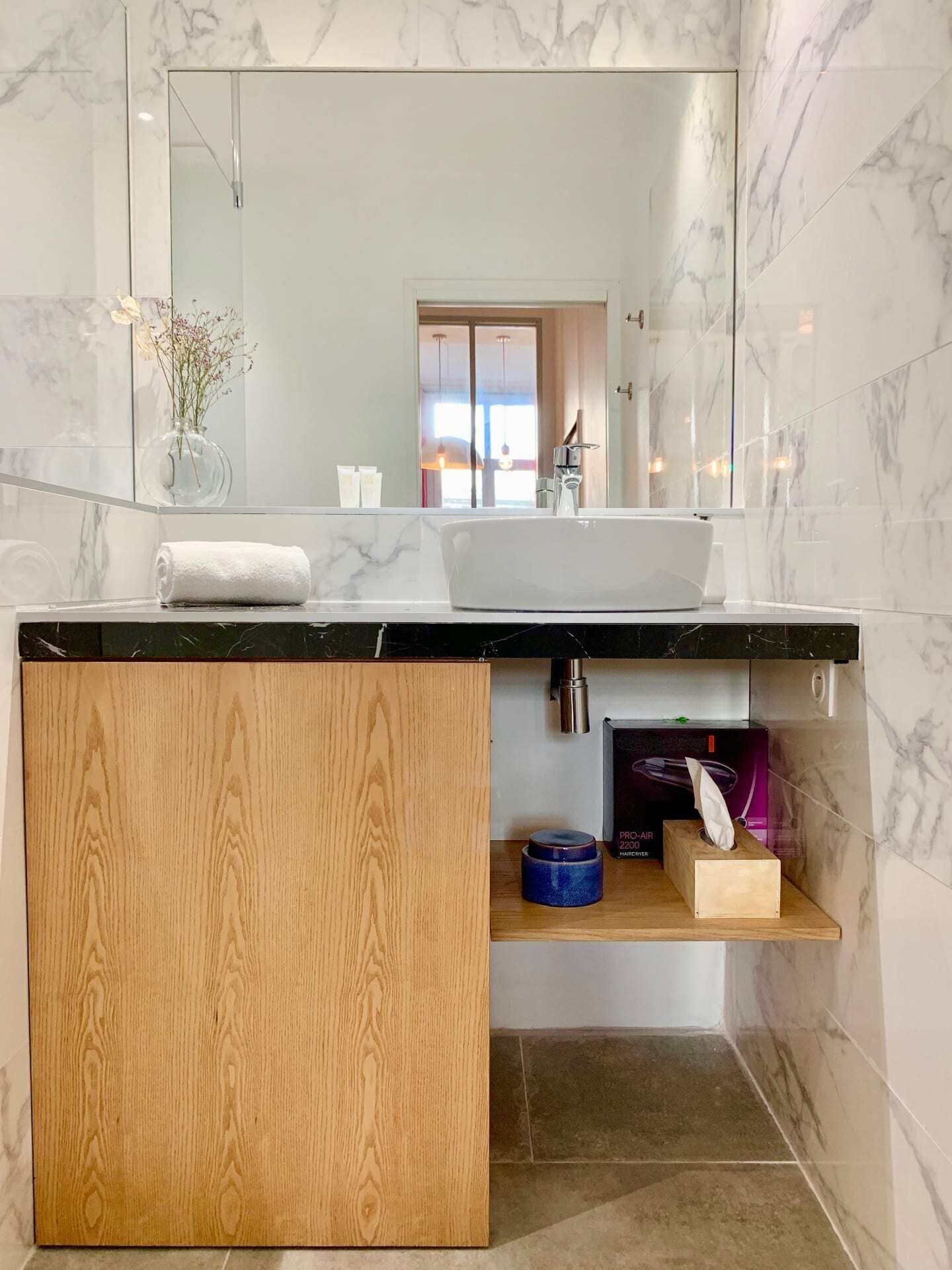 A modern bathroom with a marble sink, wooden vanity, and a mirror reflecting a door and window.