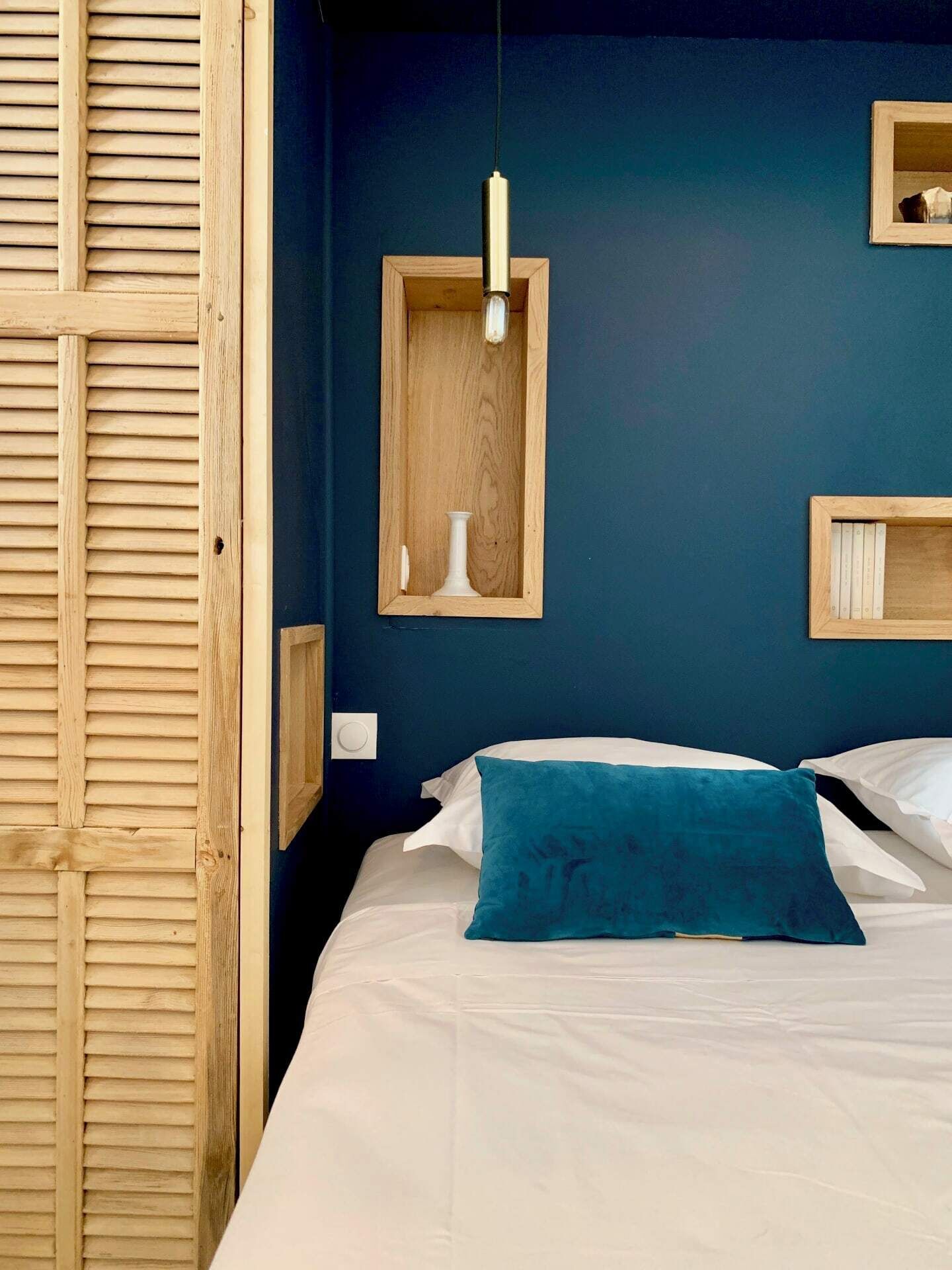 Une chambre à coucher avec un lit, un oreiller de couleur turquoise, un luminaire suspendu et des étagères en bois ornées d’objets décoratifs.