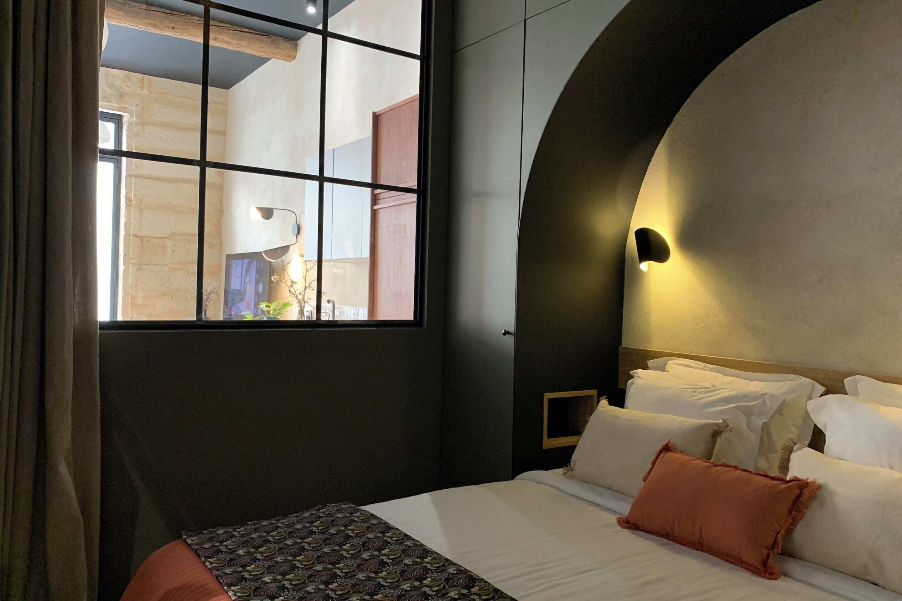 Un dormitorio con un ventanal grande, una cama con ropa de cama blanca y un plaid estampado, y una pared curva con una lámpara.