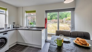 A modern kitchen with a built-in washing machine, a dining area with a table set for breakfast, and a view of a garden through large windows. - Blaen Ddol (Betws-Y-Coed)