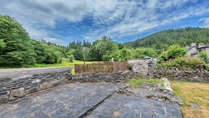 A stone pathway leading to a stone wall, surrounded by lush greenery and a forested hill in the background. - Blaen Ddol (Betws-Y-Coed)
