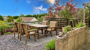 Une terrasse extérieure avec une table et des chaises en bois, entourée d’un jardin où poussent différentes plantes et délimitée par une clôture en bois.