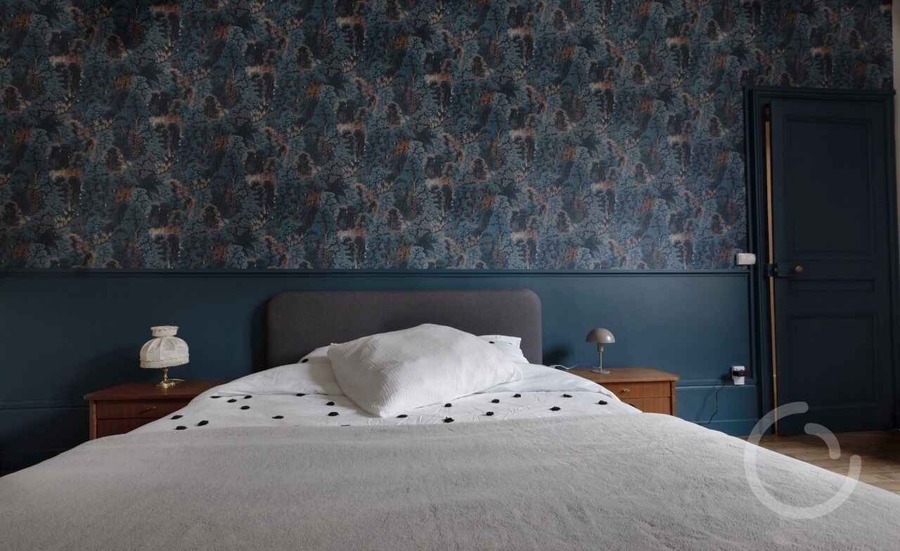 A bedroom with a patterned wallpaper, a grey headboard, a white bedspread, and two bedside tables with lamps.