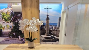Ein Empfangsschalter mit Holzoptik, ein Topf mit weißen Blüten und Blick auf eine Küstenstadt durch eine Glastür.