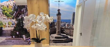 A reception desk with a wooden finish, a potted plant with white flowers, and a view of a coastal town through a glass door.