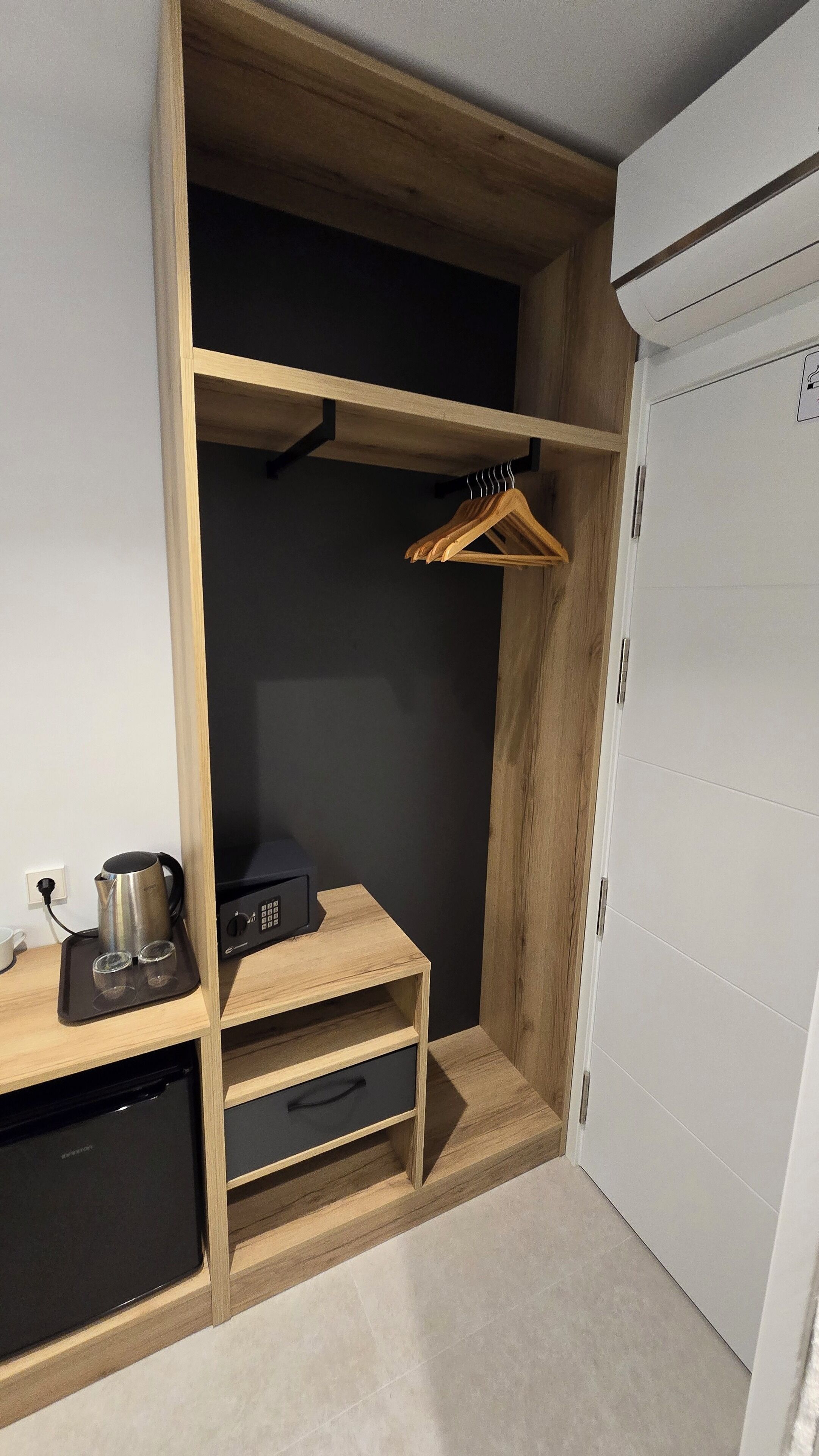 A compact room with a wooden wardrobe, a small shelf with a kettle and glasses, and a hanging clothes rack.