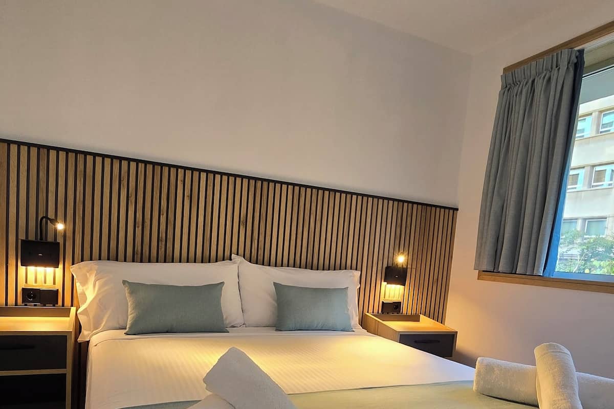 Una habitación de hotel con una cama grande, dos mesitas de noche, un cabecero de madera y una ventana con cortinas.