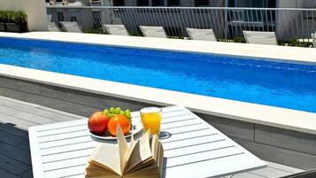 Et område ved poolen med et bord dækket med bøger, frugt og et glas appelsinjuice.