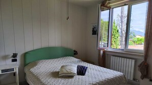 A bedroom with a green headboard, a quilted bedspread, a bedside table, and a window with a view of trees and mountains. - Your nest in Manerba (manerba del garda)