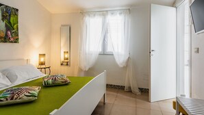 A bedroom with a bed, a nightstand, a lamp, a window with sheer curtains, and a door. - Galatea 4 by Wonderful Italy (Avola)
