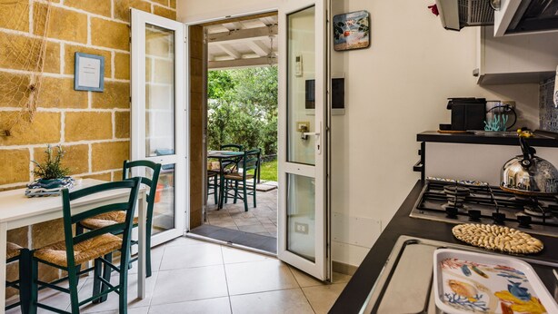 Uma cozinha com fogão a gás, mesa de jantar com cadeiras e vista para o exterior através de uma porta aberta.