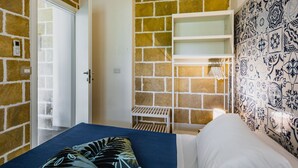Quarto com parede de tijolos, cama com colcha azul, travesseiro branco e mesa de cabeceira branca, além de parede estampada.