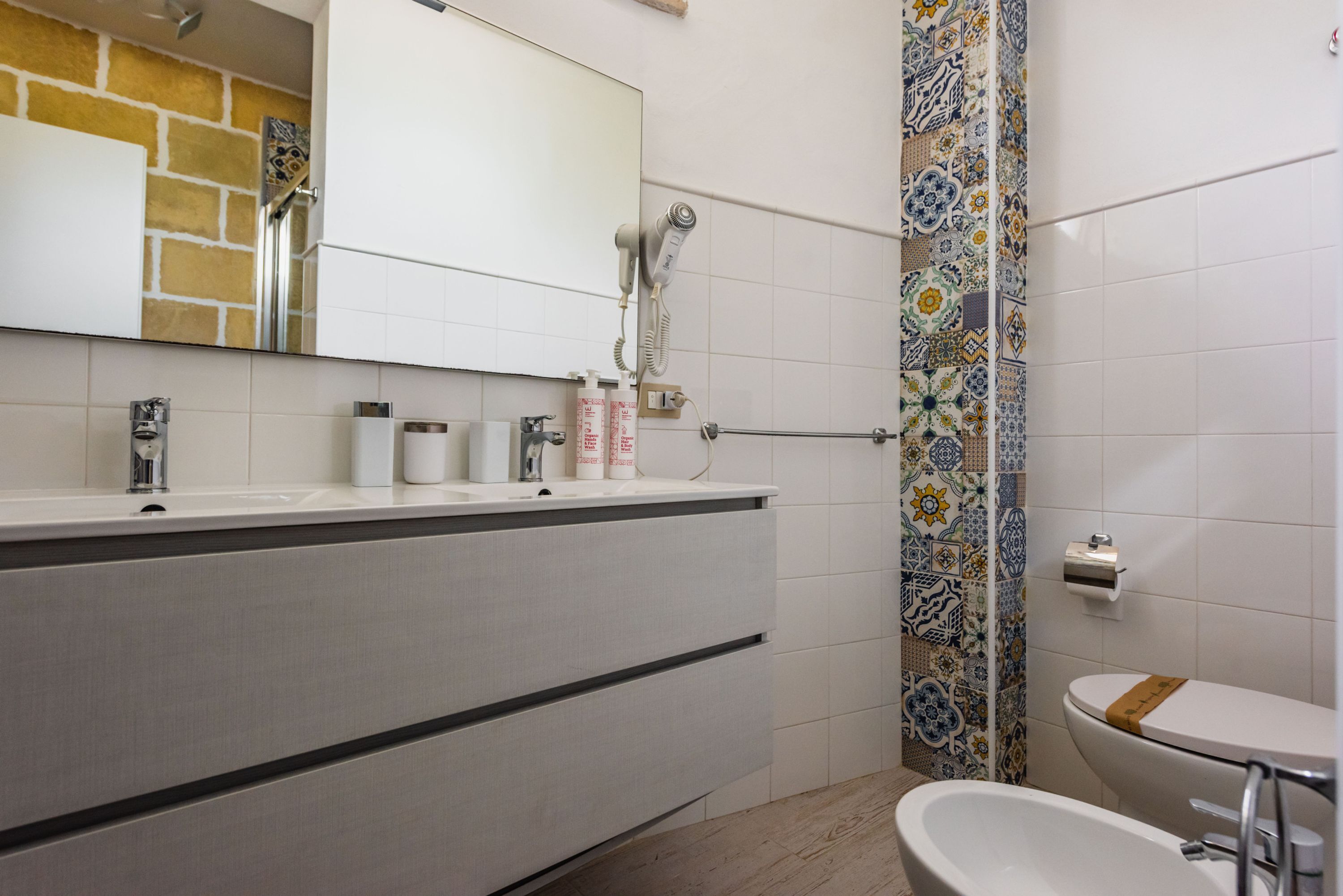 Un baño con un lavamanos blanco, espejo e inodoro. Hay un perchero y una ducha con pared de azulejos decorativos.