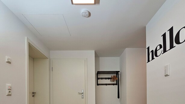 Ein Flur mit Holzboden, einem schwarz-beigen Teppich, einem an der Wand befestigten Schuhregal und einem Schild mit der Aufschrift 'hello'.