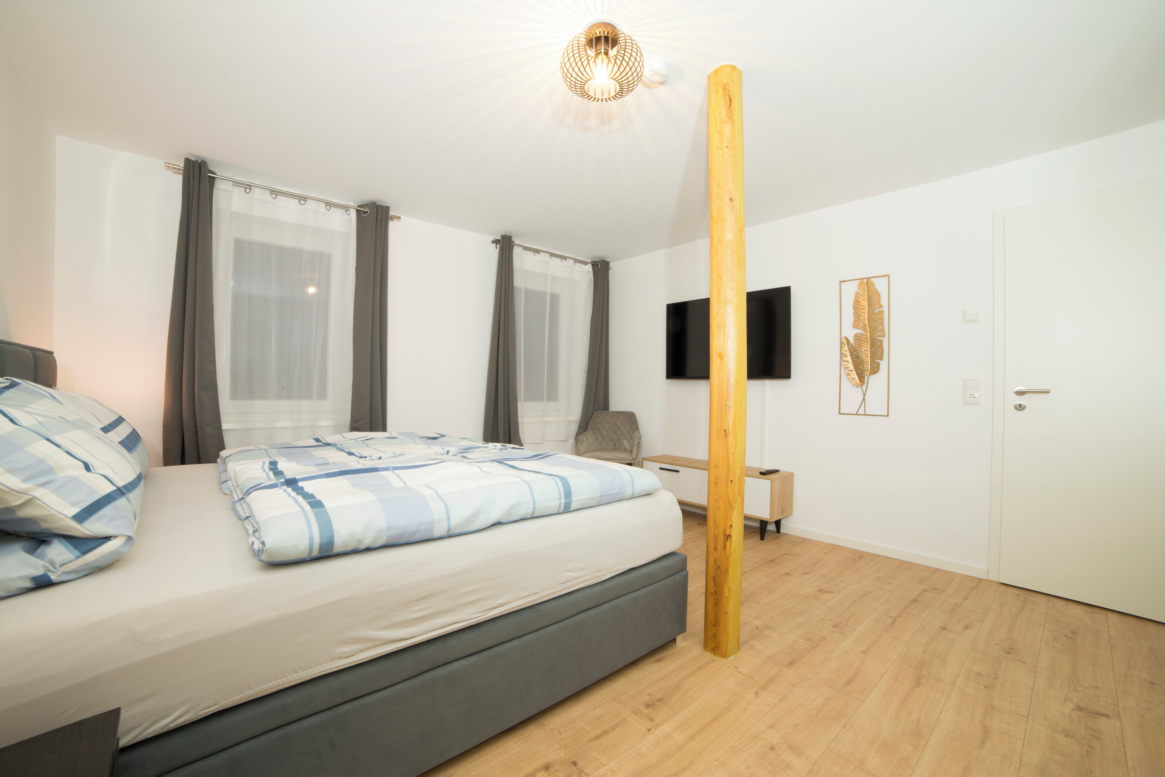 Ein modernes Schlafzimmer mit Bett, Vorhängen, einem an der Wand befestigten Fernseher und einem Holzbalken.