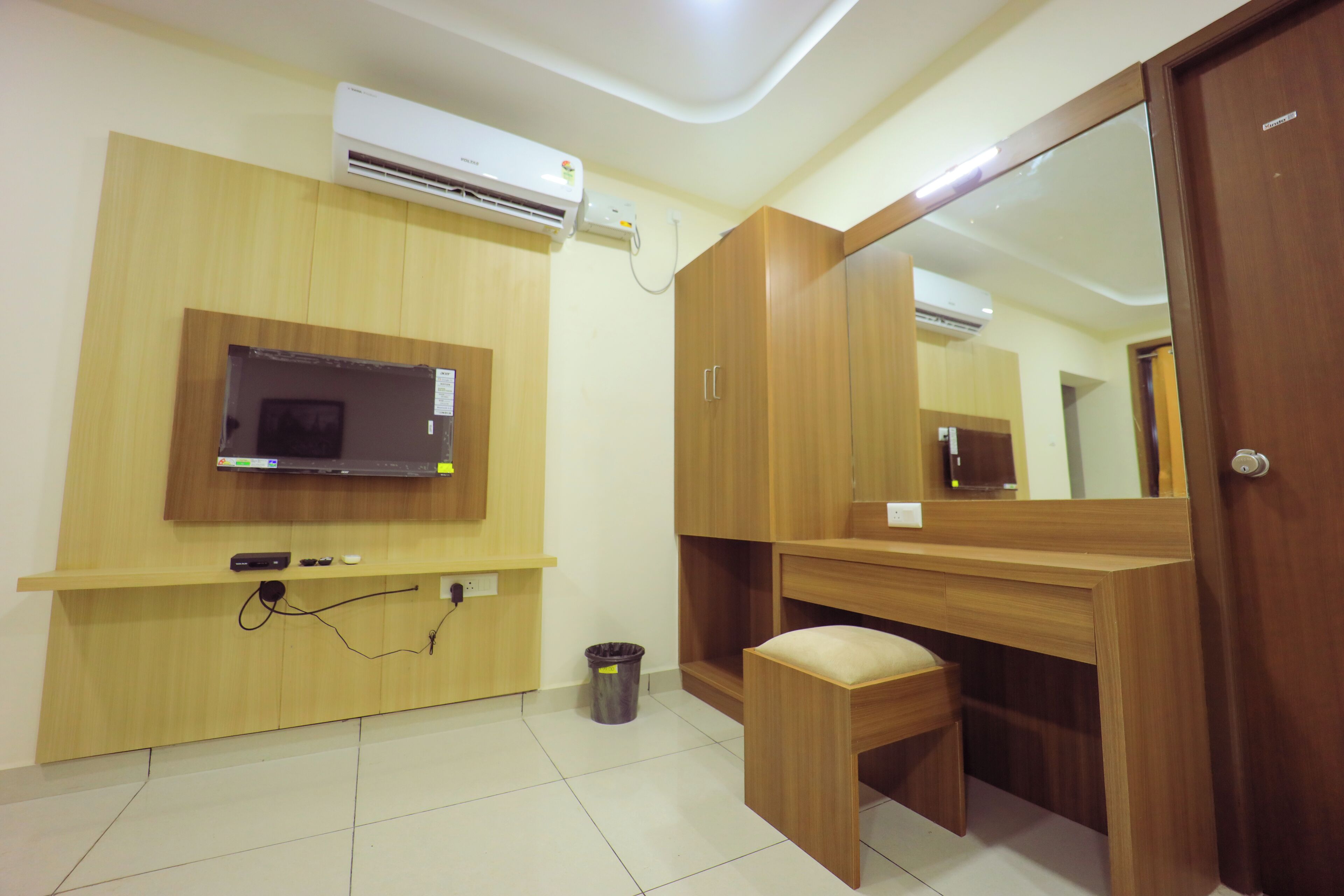 A room with a wooden desk, a television mounted on the wall, a small trash bin, and a mirror.