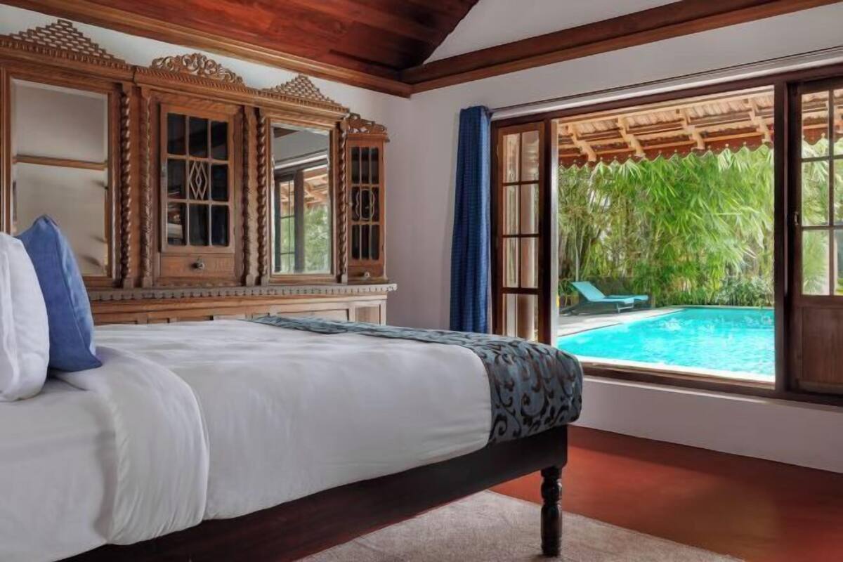 A bedroom with a large bed, wooden furniture, a view of a swimming pool, and a window with a view of greenery.