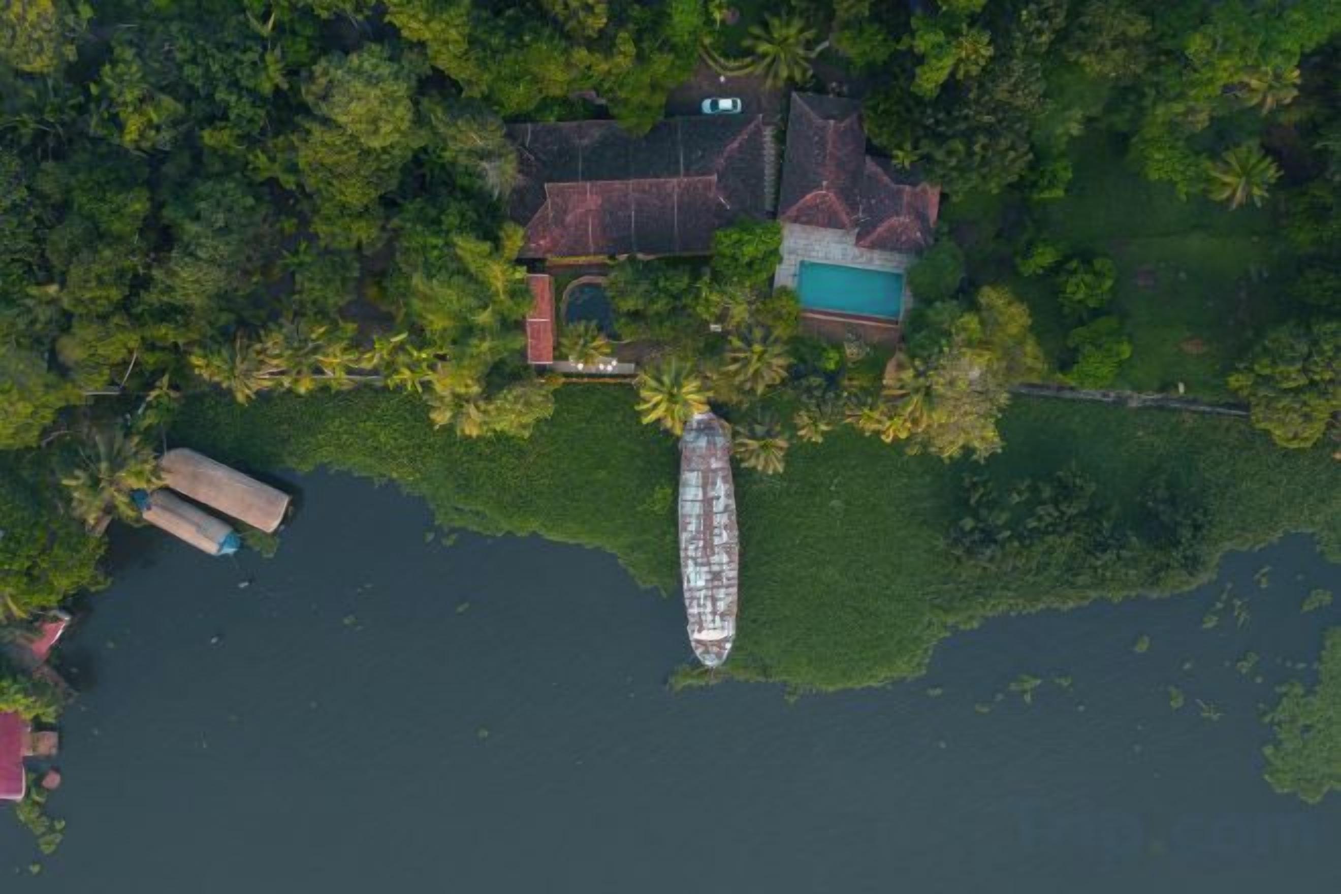 An aerial view of a house with a swimming pool, surrounded by dense greenery and a dock.