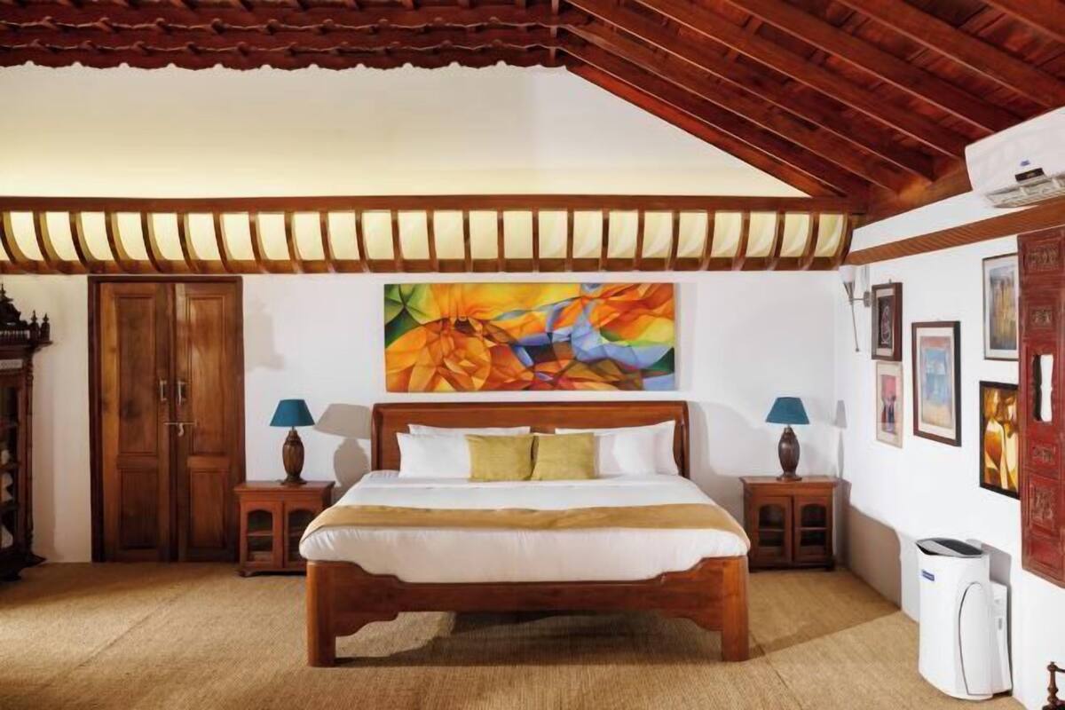 A bedroom with a wooden bed, two bedside tables with lamps, a large abstract painting on the wall, and a ceiling with wooden beams.