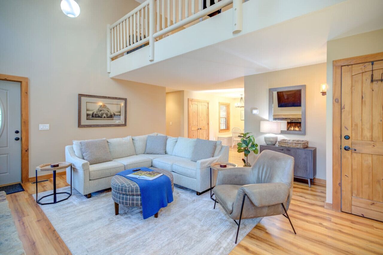 A spacious living room with a large L-shaped sectional sofa, a small side table, and a chair. A wooden door and a staircase are visible in the background.