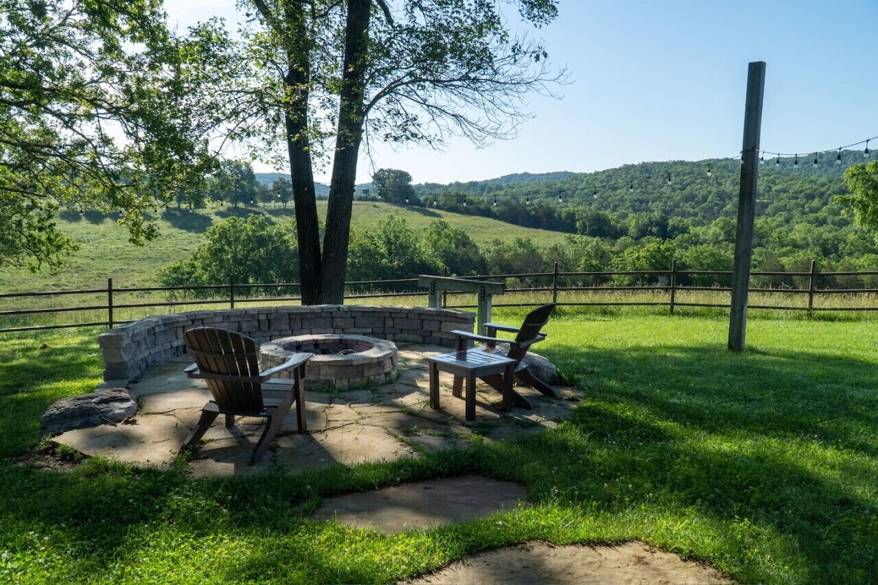 Un espace extérieur aménagé avec un foyer en pierre, deux fauteuils Adirondack et une vue sur une colline verdoyante.