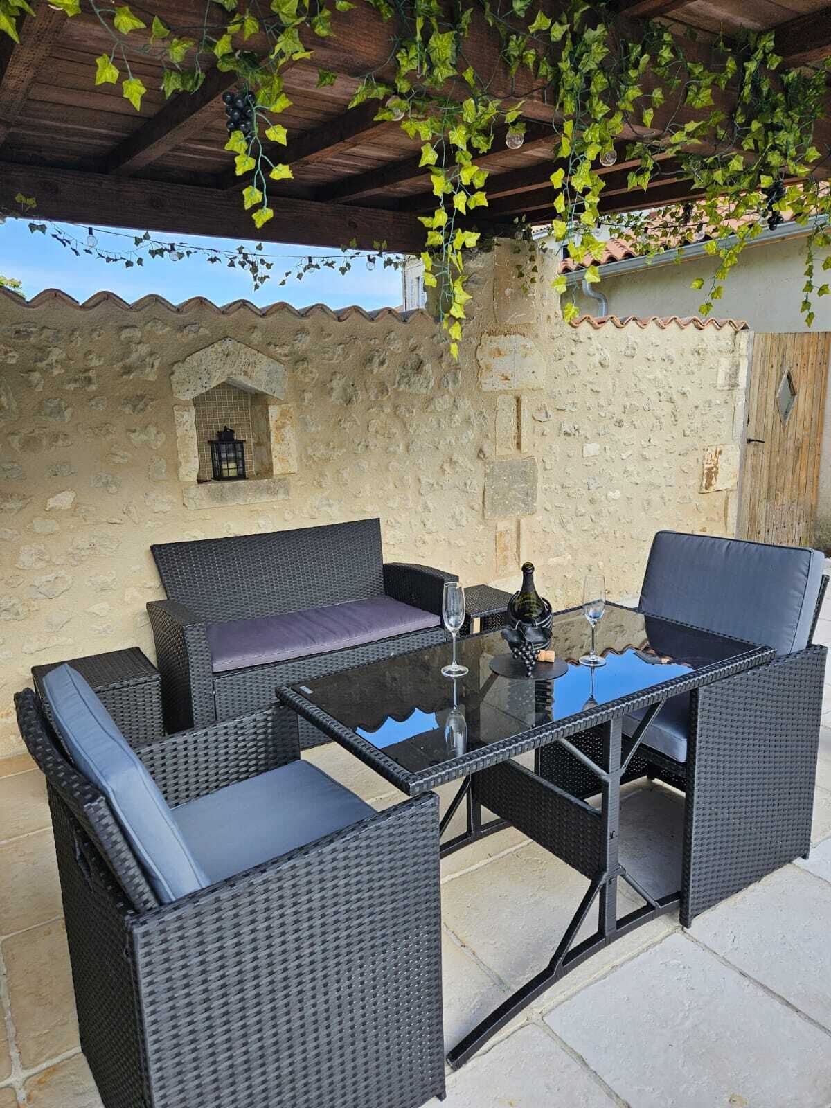 Un área de estar al aire libre con mobiliario de mimbre y una mesa de vidrio, frente a una pared de piedra con techo de tejas.