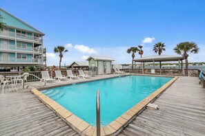 A pool area with a wooden deck, lounge chairs, and a building with a balcony.