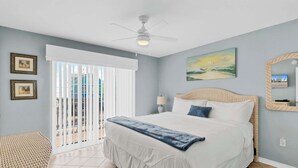 A bedroom with a large bed, a ceiling fan, a balcony with white blinds, and a beach-themed painting on the wall. - Waterfront Gulf Shores Condo w/ Beach Access (Gulf Shores)
