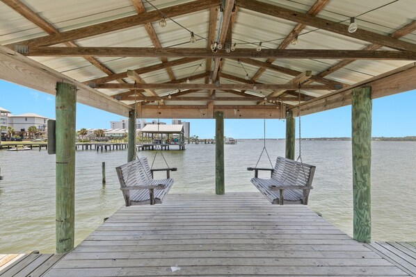 A wooden pier with two benches under a covered area, overlooking a body of water with buildings in the background.