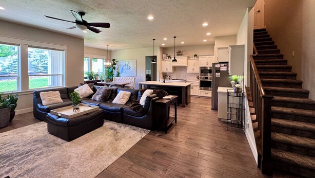 A spacious living room with a dark leather sofa, a wooden coffee table, and a large window overlooking a garden. A kitchen with white cabinets and stainless steel appliances is visible in the background.