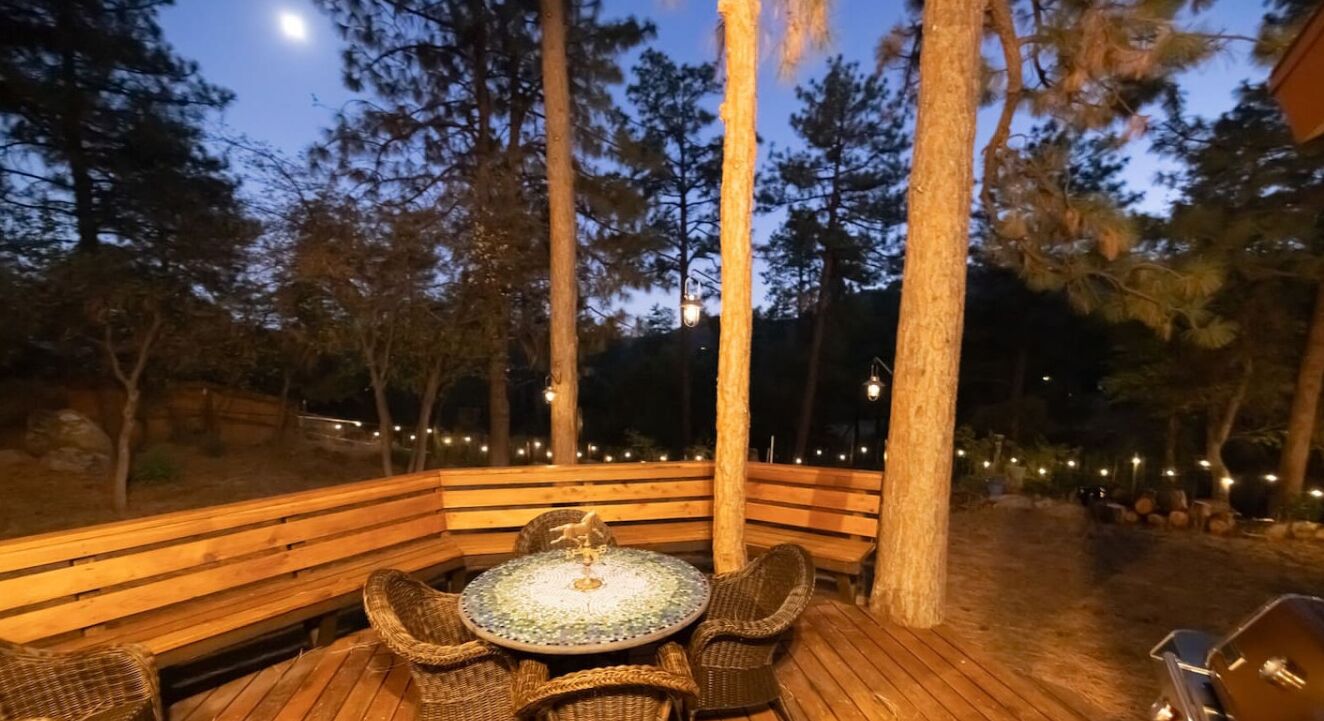 An outdoor seating area with a wooden bench and wicker chairs, surrounded by tall trees and string lights.