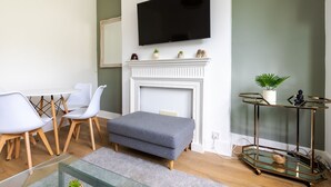 Ett modernt vardagsrum med en vit öppen spis, en väggmonterad TV, ett soffbord i glas och en sittgrupp med fåtöljer och en soffa.