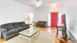A living room with a gray sofa, a glass coffee table, a patterned armchair, a TV on a stand, and a red door in the background.