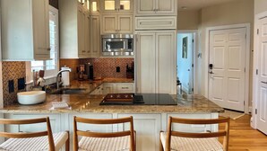A modern kitchen with white cabinetry, a large island with bar stools, a built-in microwave, and a patterned backsplash. - Modern beachfront condo with private deck, harbor views, and parking! (Provincetown)