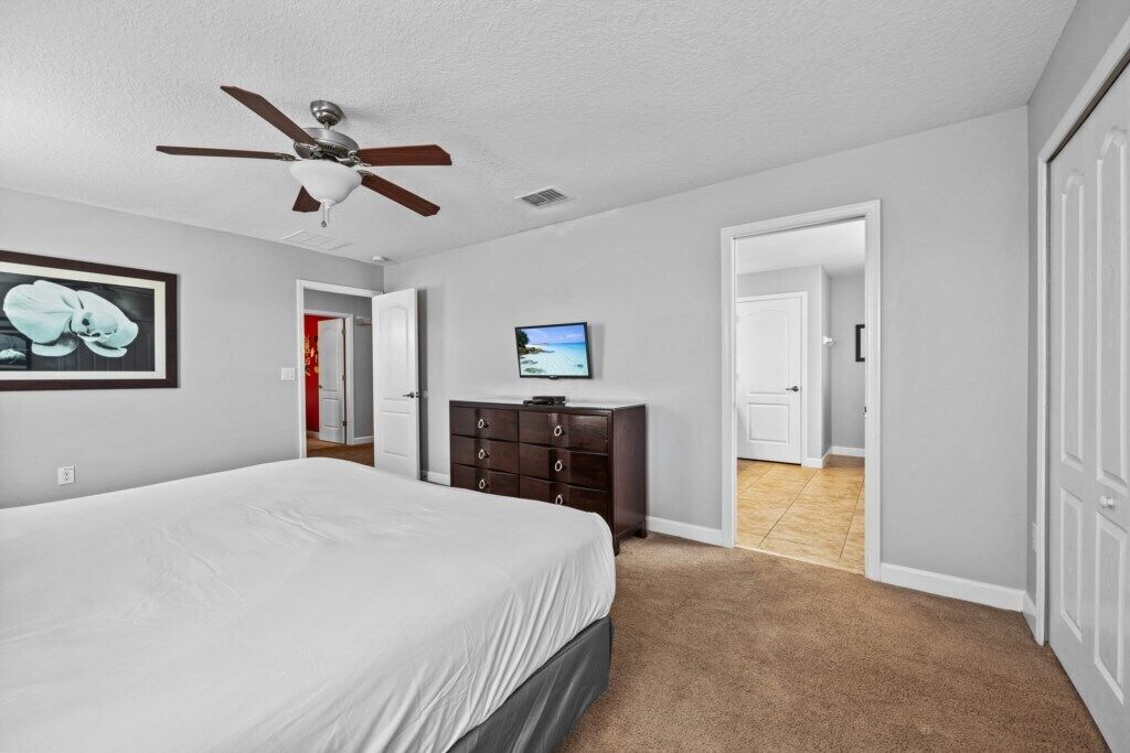 A bedroom with a bed, a ceiling fan, a dresser, a TV, and a door leading to another room.