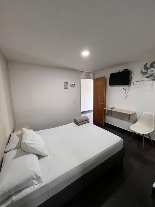 A modern hotel room with a bed, a desk, a chair, a TV, and a door. - AURA (Medellín)