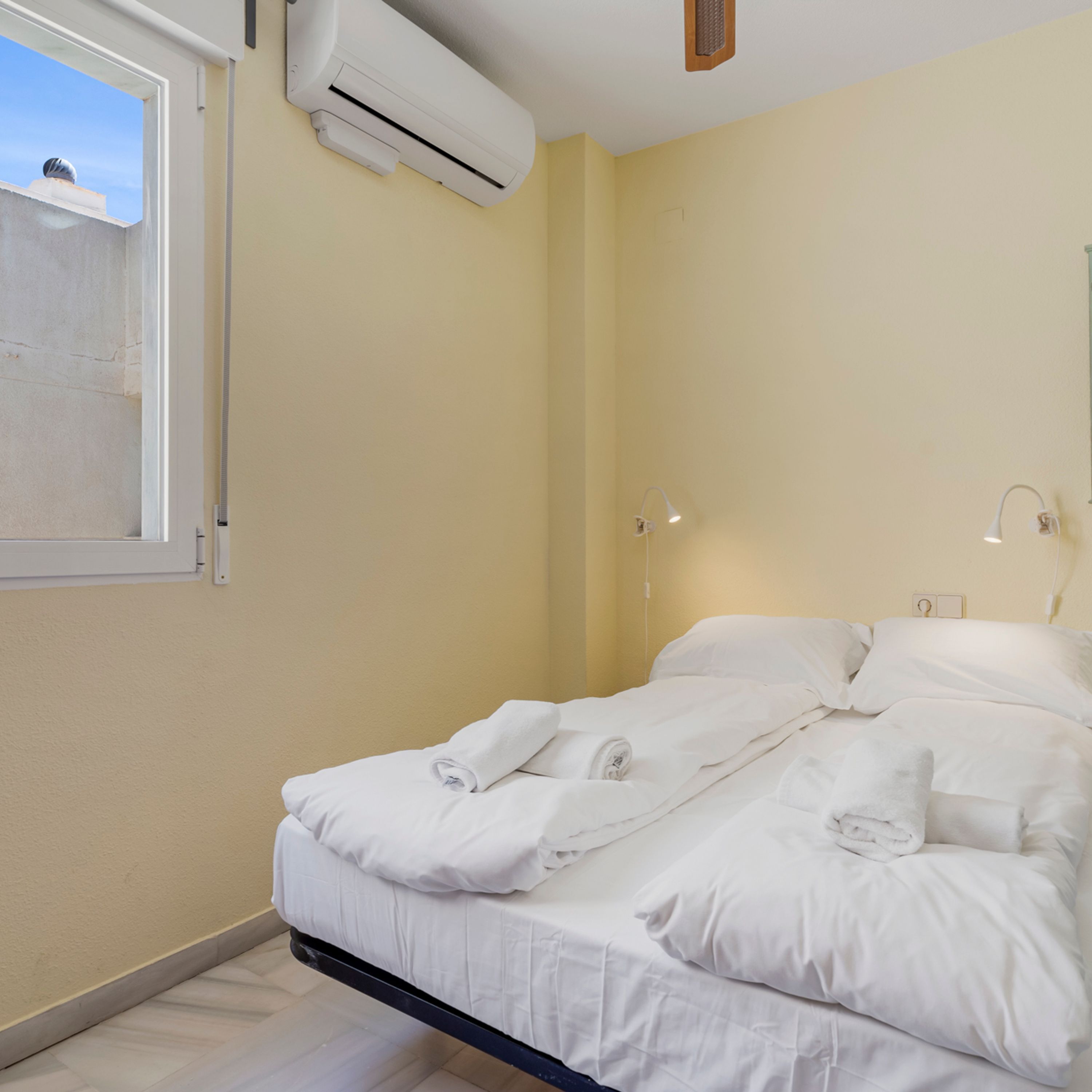 A bedroom with a bed, white linens, an air conditioning unit, a window, and a wall-mounted light fixture.