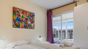 A hotel room with a large bed, a nightstand, a painting on the wall, and a window with a view of buildings. - Los Boliches Sunny Apartment - Ref 120 (Fuengirola)