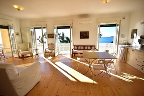 A spacious living room with a wooden floor, a sofa, a dining table, and chairs. There is a balcony with a view of the sea. - Panorama Apartment in Ospedaletti by Wonderful Italy (Ospedaletti)