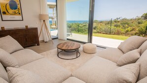 A modern living room with a large sofa, a coffee table, and a view of the ocean. - Villa 142 Bahia de las Rocas (Manilva)