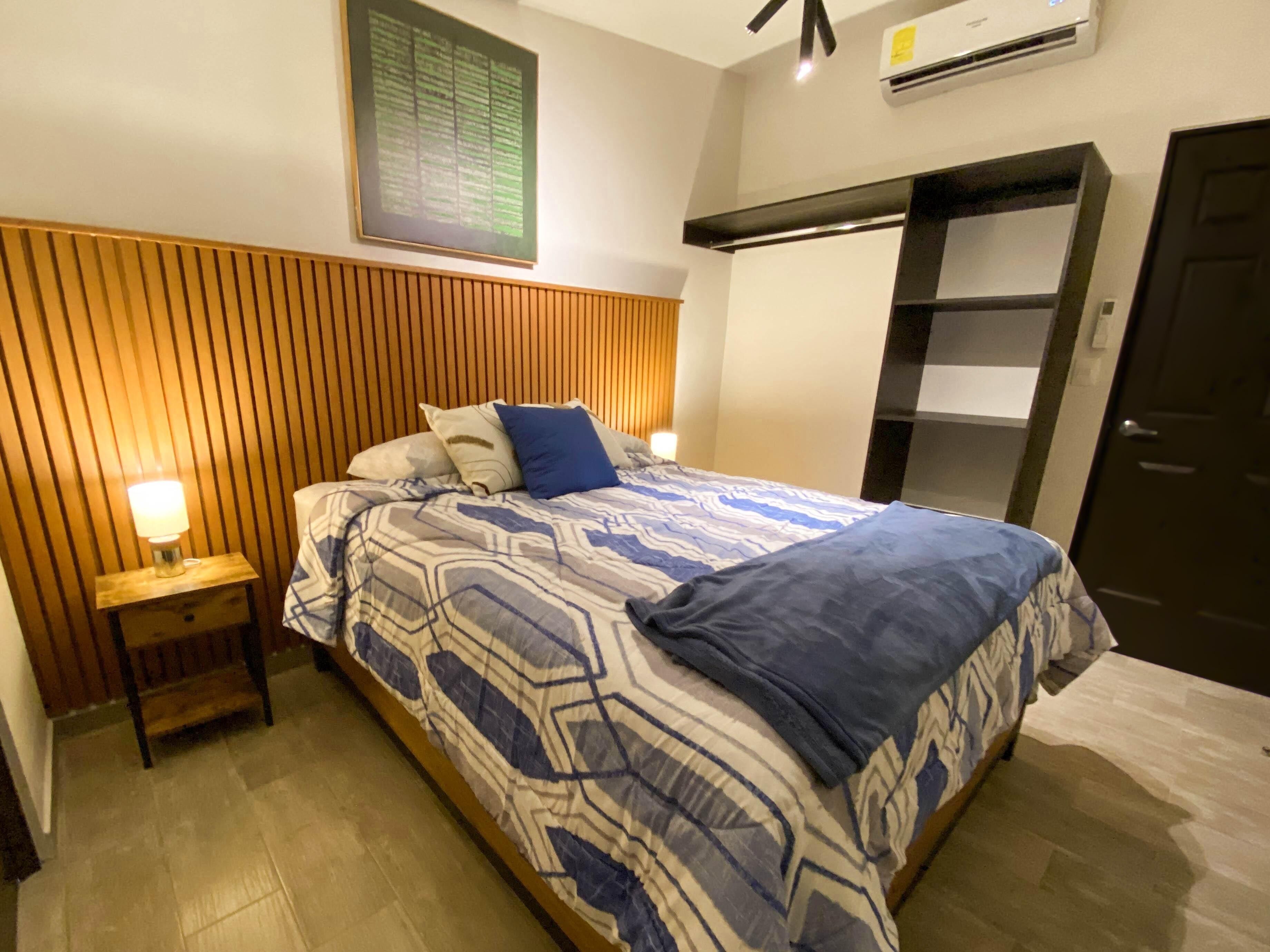 Una habitación de hotel con cama, mesita de noche, aire acondicionado de pared y una puerta.