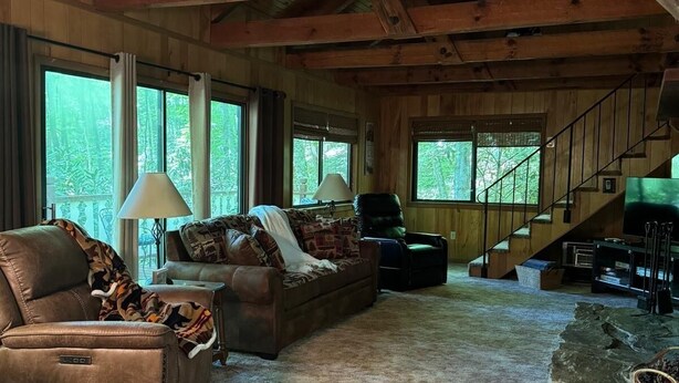 A spacious living room with a high ceiling, wooden beams, a fireplace, a staircase, and large windows offering a view of the outdoors. - My Cabin In The Mountain (Otto)