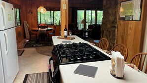 A rustic cabin interior with a kitchen featuring a gas stove, a dining area with a table and chairs, and large windows offering a view of the forest. - My Cabin In The Mountain (Otto)