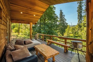 Une terrasse en bois avec un canapé, des coussins et une table, donnant sur une zone boisée.